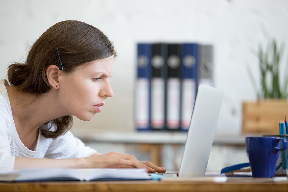 Jovem mulher branca com cabelos castanhos. Ela está com rosto próximo à tela do laptop, com dificuldades para enxergar.