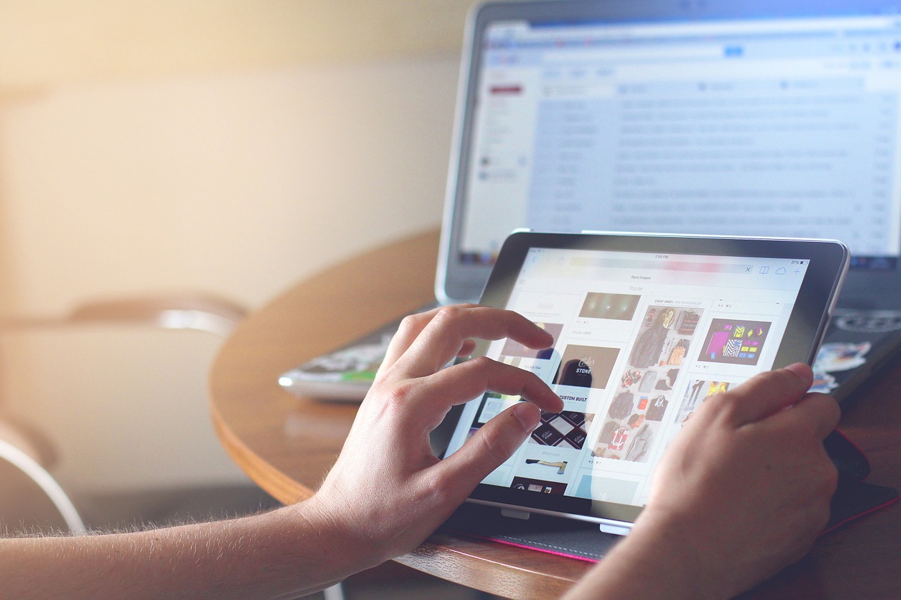 Foto de uma pessoa segurando um Ipad e tocando a tela com uma das mãos. Ao fundo, há um laptop aberto. Os dois aparelhos estão em cima de uma mesa.