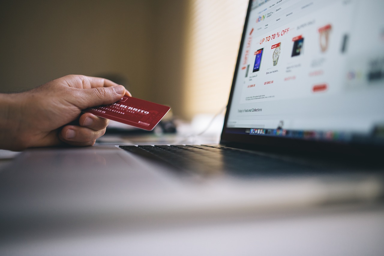 Foto de uma mão segurando um cartão de crédito em frente a um laptop. Na tela, há fotos de relógio e outros produtos que estão desfocados.