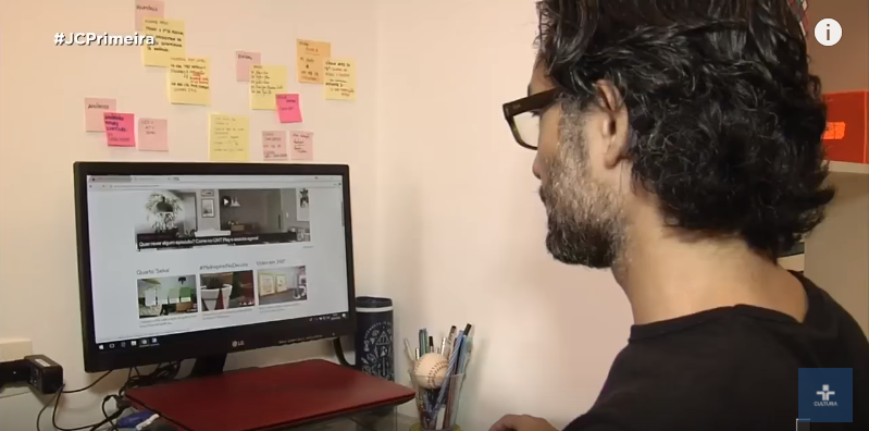 Homem de óculos olhando para a tela de um computador em uma sala. Há diversos post-its coloridos colados na parede. 