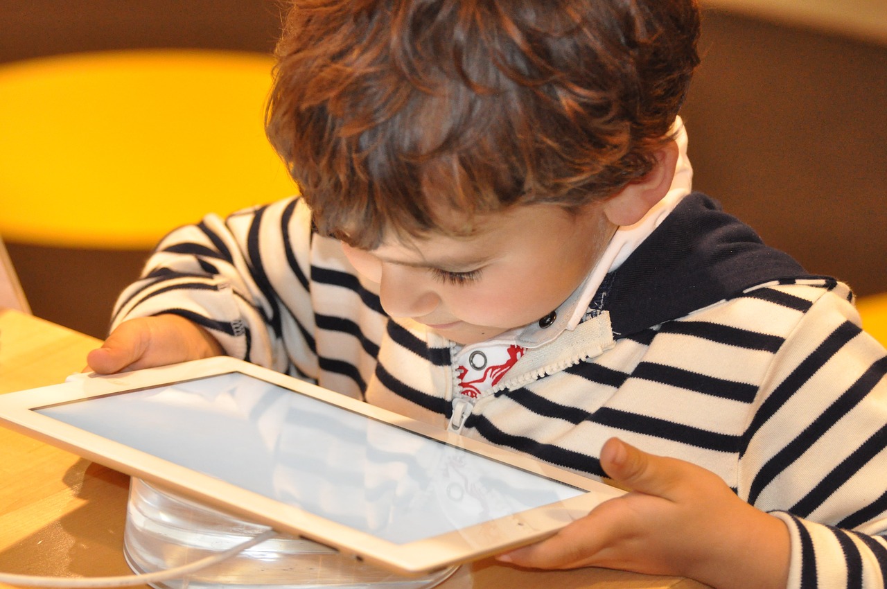 Foto de uma criança segurando um tablet com as duas mãos. Ela sorri e está com o rosto muito próximo à tela.