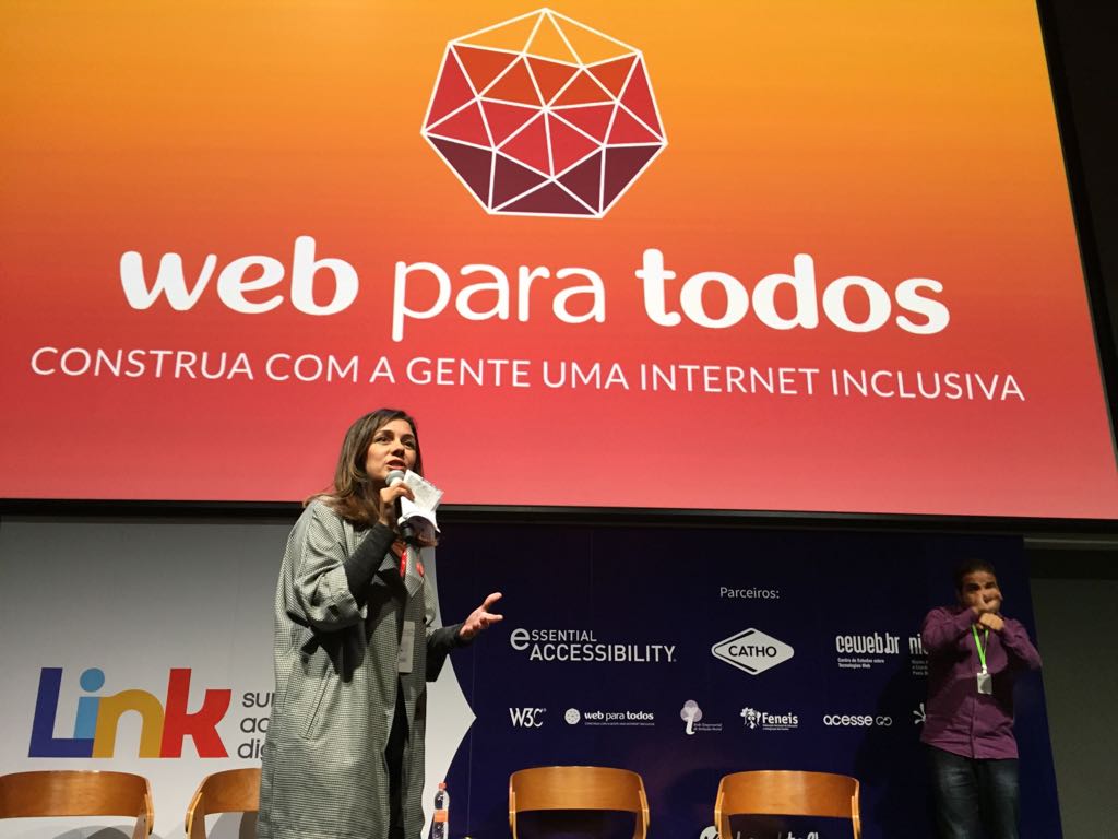 Foto da Simone Freire no palco do Google Campus. Ela está pé, falando ao microfone, olhando para o público.