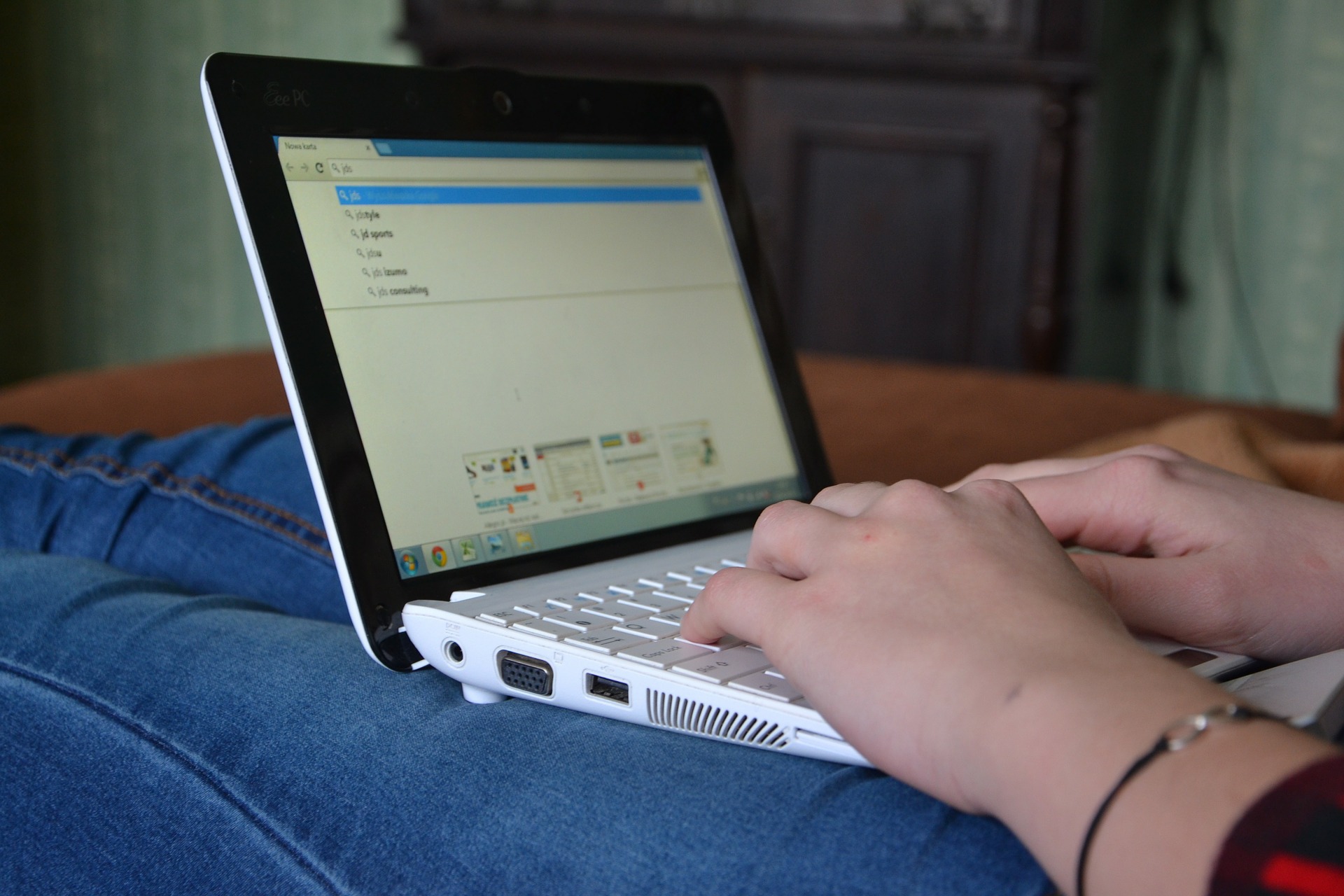 Pessoa sentada com um laptop nas pernas e com as mãos no teclado do equipamento. A tela mostra diversas palavras na ferramenta de busca do navegador.