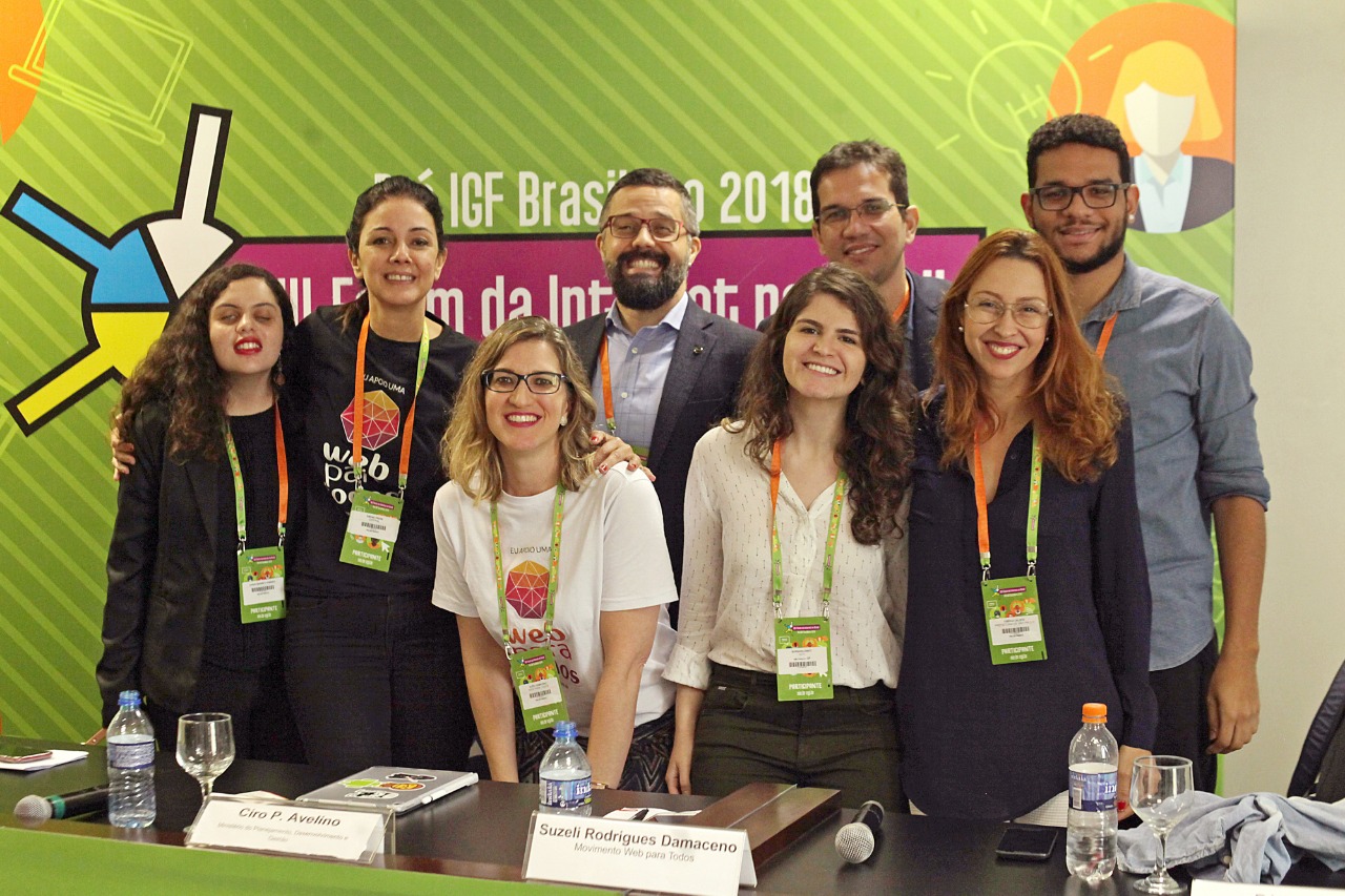 Foto dos oito integrantes do painel, em pé, sorrindo atrás de uma bancada e abraçadas. Ao fundo, há um painel com o texto: "VIII Fórum da Internet no Brasil".