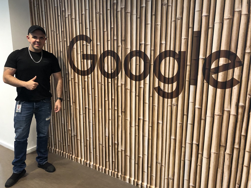 Leandro Pólito está em pé, sorrindo, ao lado de um painel com o nome Google. Ele faz sinal de positivo com uma das mãos. 