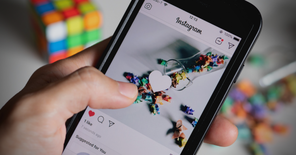 Foto da mão de uma pessoa que segura um celular. A tela mostra o aplicativo Instagram com uma foto de objetos coloridos. Há um ícone de coração no centro da foto. Ao fundo, há um cubo mágico. 
