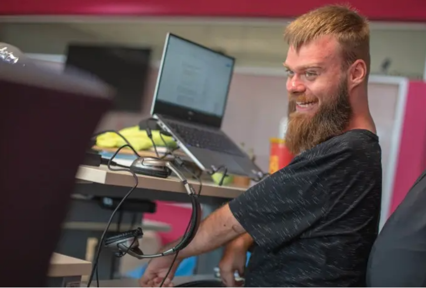 Foto de um homem ruivo com deficiência nos braços e nas mãos sorrindo, sentado em frente ao computador.