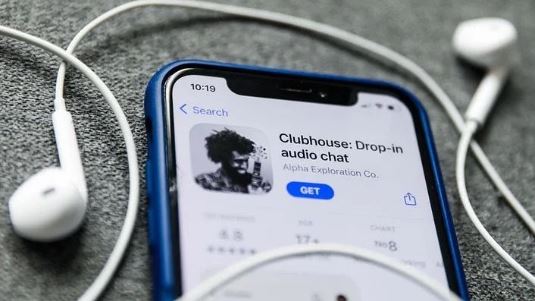 Foto em tons de cinza de um celular com bordas azuis e fones de ouvido. Na tela, há a imagem do aplicativo da Clubhouse com o botão "get" destacado em azul.