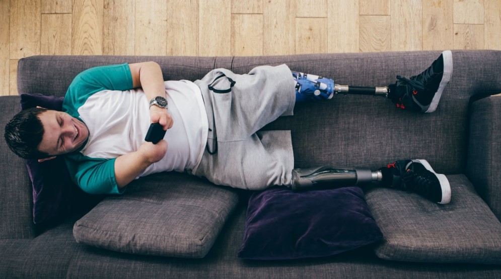 Foto de um jovem branco deitado em um sofá. Ele usa próteses nas duas pernas e tem os dois braços atrofiados. Ele segura um celular com as duas mãos, olha para a tela e sorri.
