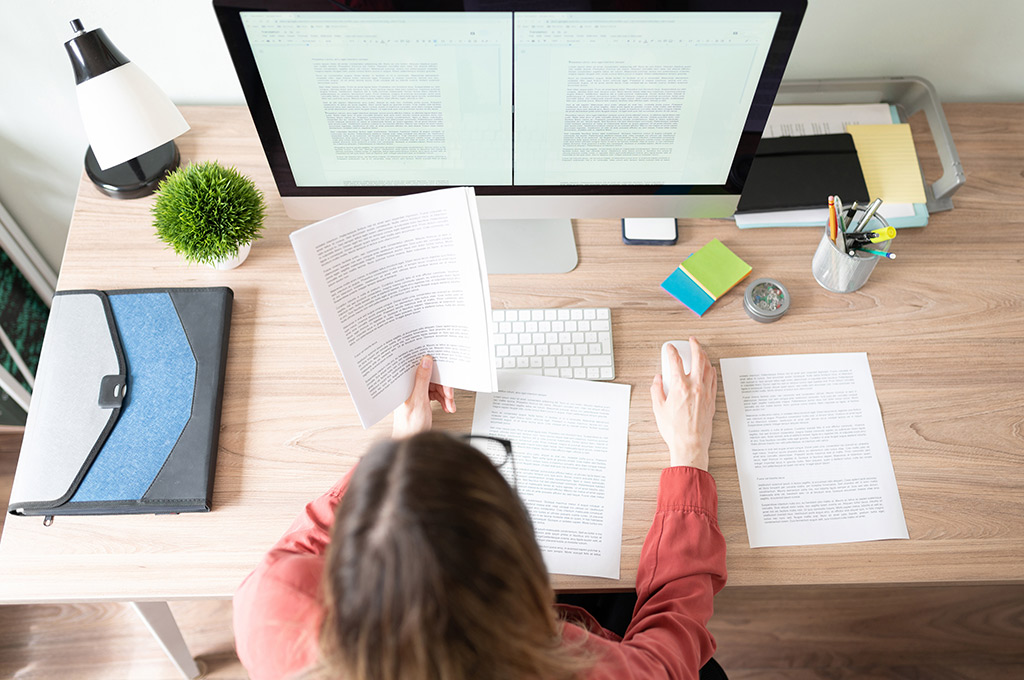 Foto tirada de cima de uma mulher de costas mexendo em um computador. O monitor mostra duas páginas de texto e ela segura diversas folhas com um texto impresso.