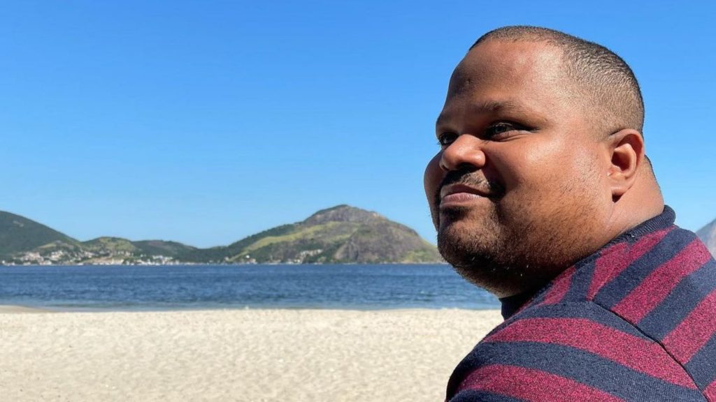 Foto de Diego Conceição de perfil e em primeiro plano. Ele é homem negro, tem olhos verdes e sorri. Está em uma praia em um dia ensolarado.