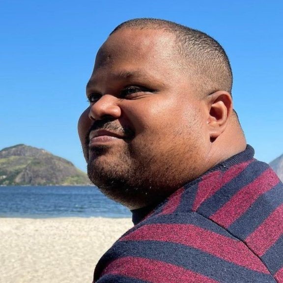 Foto de Diego Conceição de perfil e em primeiro plano. Ele é homem negro, tem olhos verdes e sorri. Está em uma praia em um dia ensolarado.