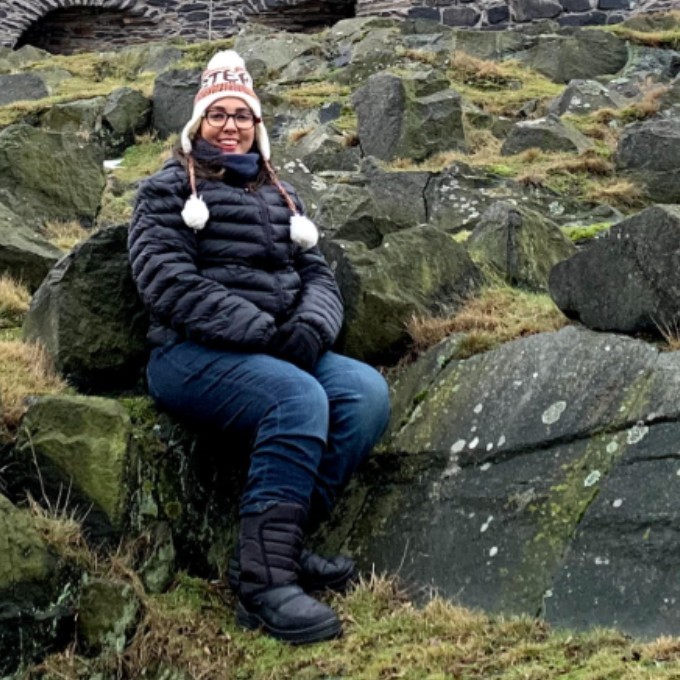 Foto de Marília Suzart sorrindo, sentada à frente de uma parede de pedras. Ela é branca, usa óculos de grau arredondado, veste jaqueta de frio, gorro, luvas e botas pretas.