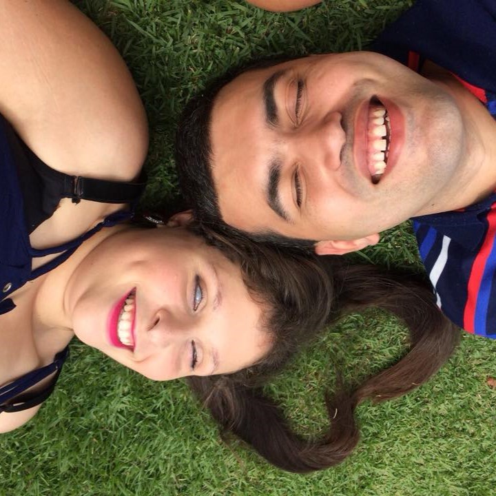 Foto de Leonardo Gleison e Camila Domingues deitados na grama em posições opostas e com suas cabeças lado a lado. Os cabelos da Camila estão ajeitados em formato de coração sobre a grama. Os dois sorriem de olhos fechados.