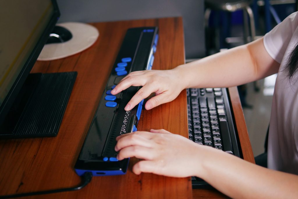 Foto de uma régua Braille em destaque tocada pelas mãos de uma pessoa. O equipamento está sobre uma mesa com teclado, mouse e monitor de computador.