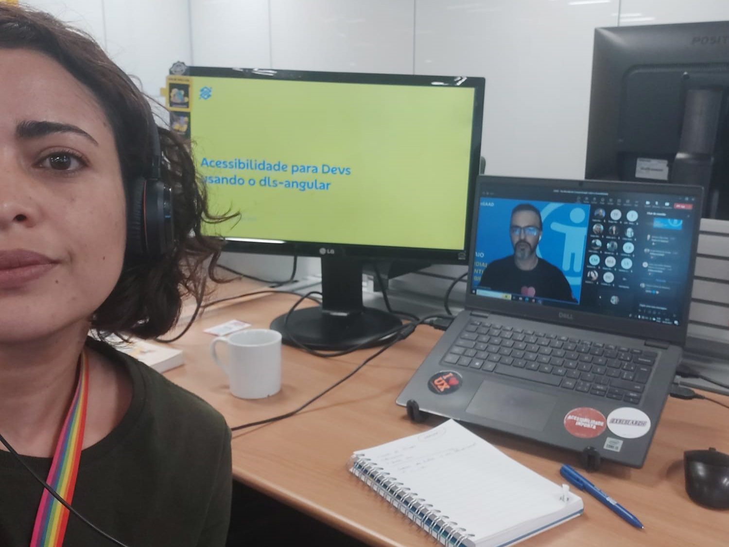 Foto de Marília Rodrigues em frente a uma mesa de escritório com um notebook, que tem na tela a projeção de uma reunião virtual com várias pessoas, e ao lado há um monitor grande com o texto "Acessibilidade para Devs usando o dis-angular". 