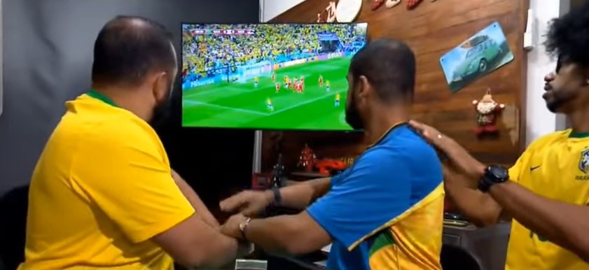 Foto de torcedor surdocego que assiste a um jogo de futebol com auxilio de dois intérpretes de Libras tátil. Eles estão em uma sala com TV ao fundo que exibe uma partida da Copa.
