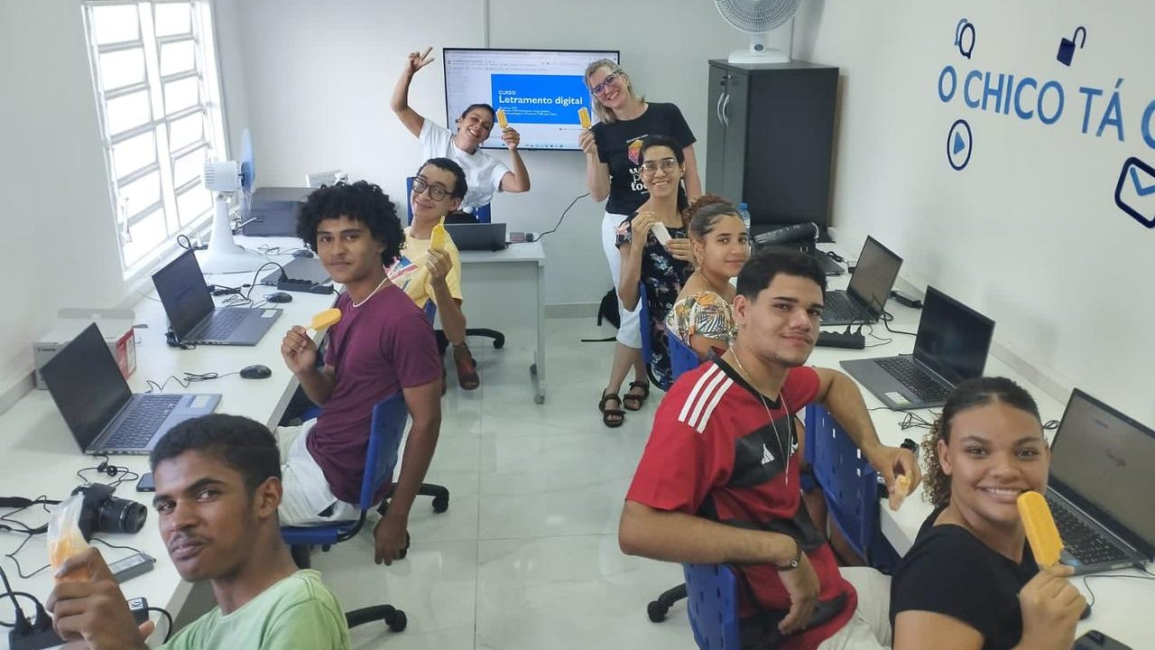 Foto de 9 pessoas dentro de uma sala de aula equipada com notebooks. Todas seguram um picolé e olham sorrindo para a câmera.