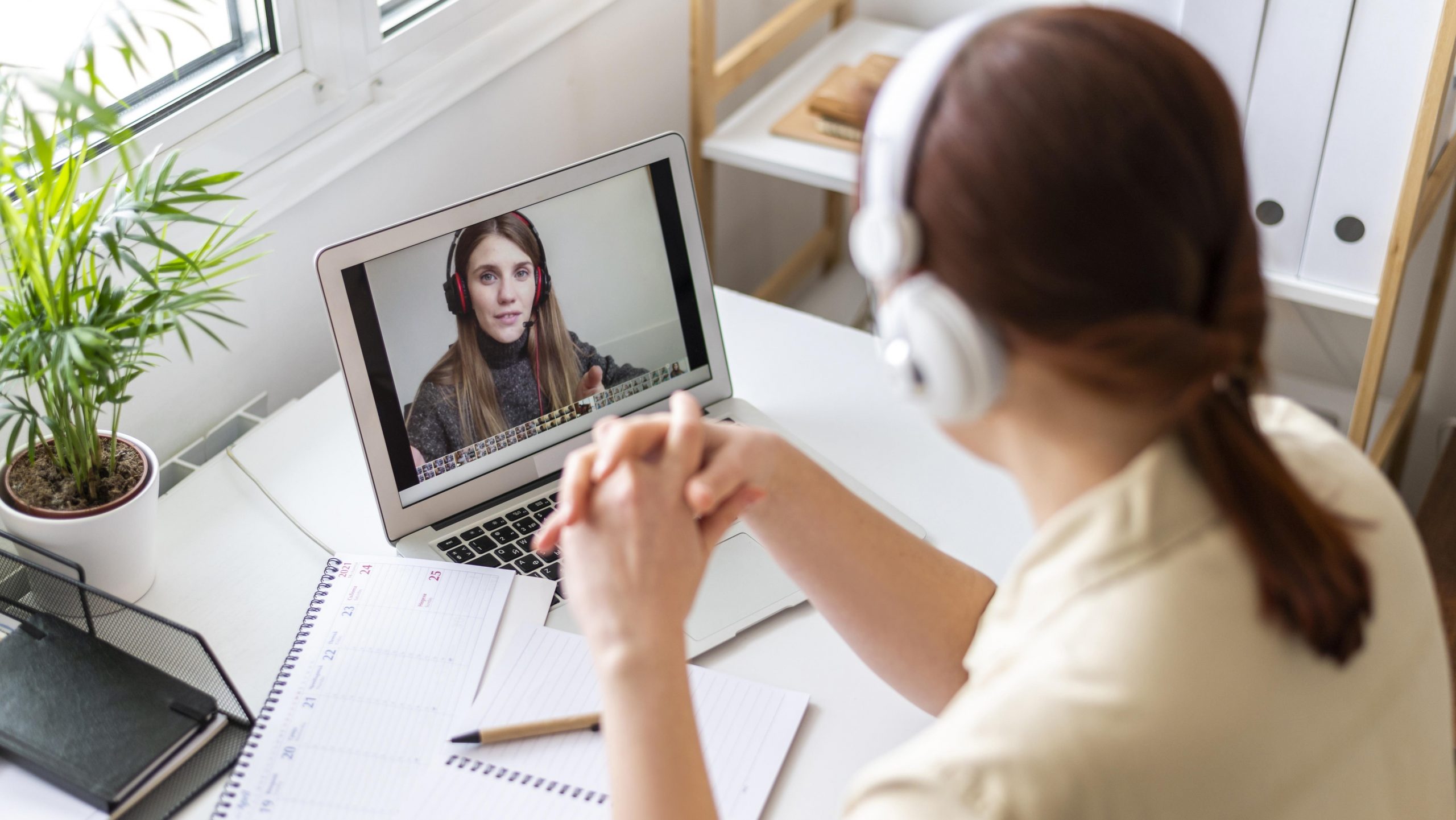 Foto de uma mulher em um escritório sentada de frente para um notebook, onde há outra mulher projetada na tela. As duas usam fones de ouvido e parecem estar em uma videochamada.