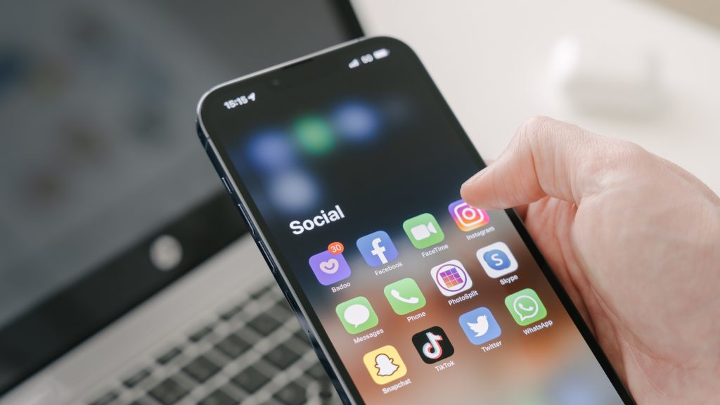 Foto de mão segurando um celular com vários ícones de aplicativos de redes sociais na tela.