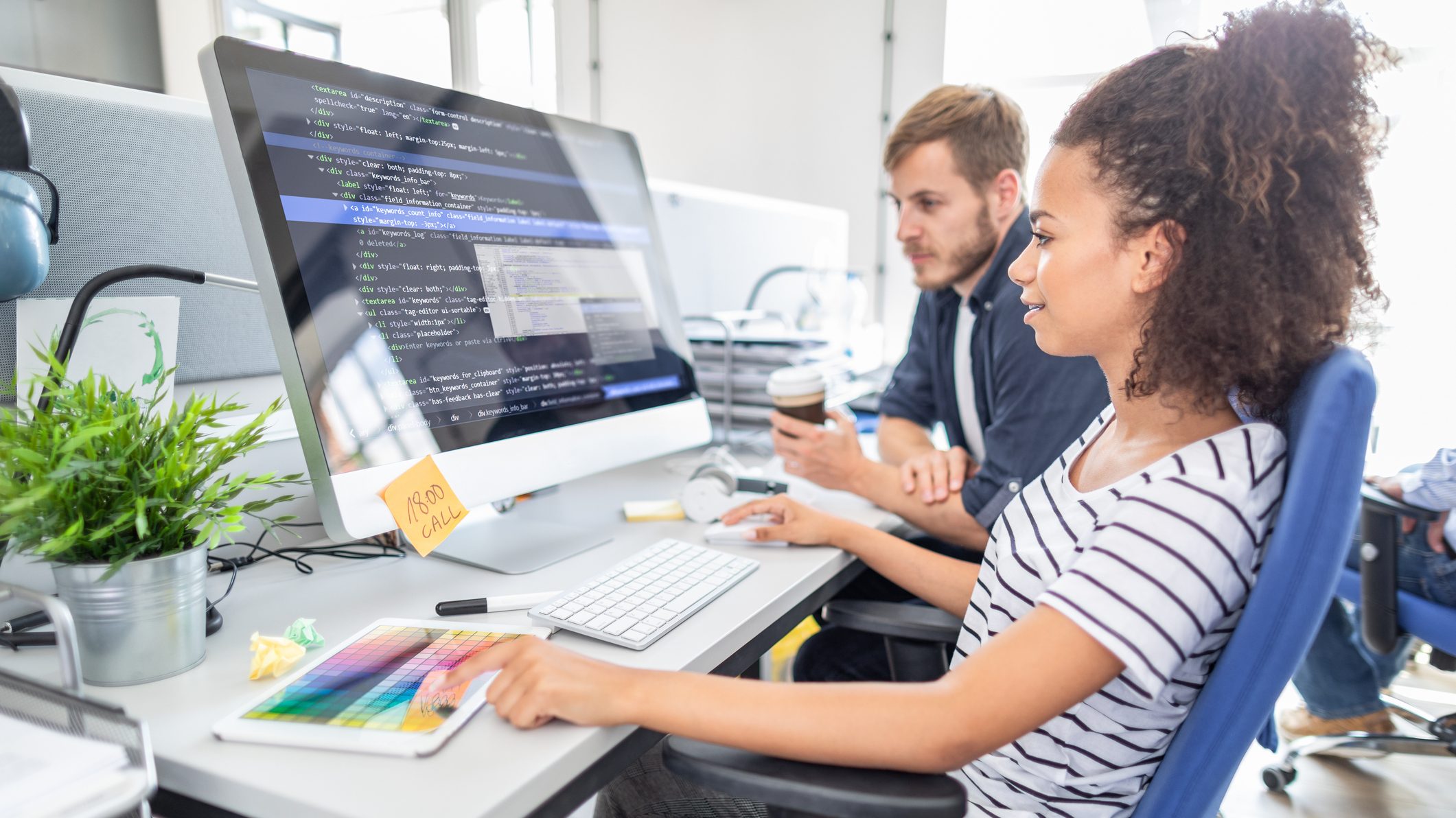 Foto de jovem negra em frente a um computador com tela grande, onde estão projetadas linhas de código. Ao lado dela há um jovem que olha para a tela enquanto ela toca no mouse.