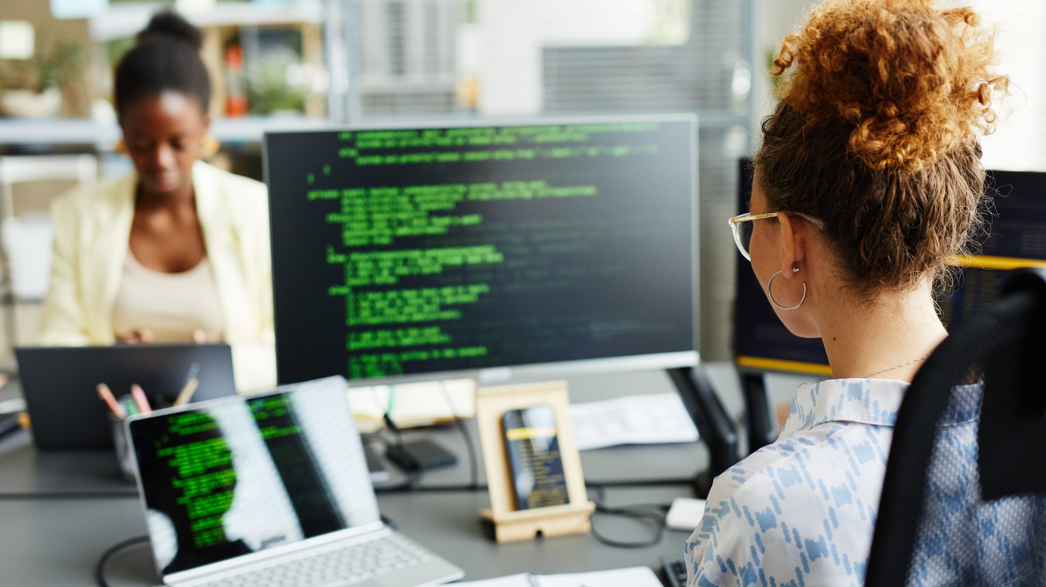 Foto de duas mulheres digitando em computadores. Uma delas está em frente a três telas com códigos de programação.