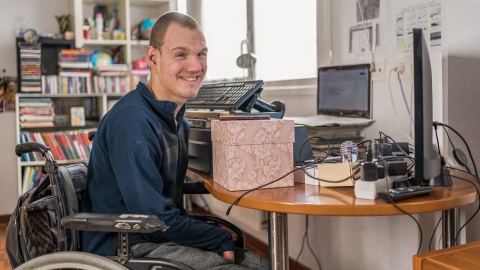 Foto de um jovem com deficiência motora em uma cadeira de rodas. Ele está num ambiente fechado, em frente a uma mesa com computadores, tecnologias assistivas, entre outros objetos.