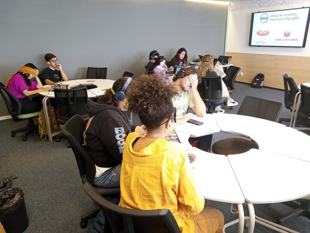 Foto da sala de sala de aula com três grandes mesas redondas. Ao redor delas há estudantes com dispositivos eletrônicos, fones de ouvido e olhos vendados ou fechados.