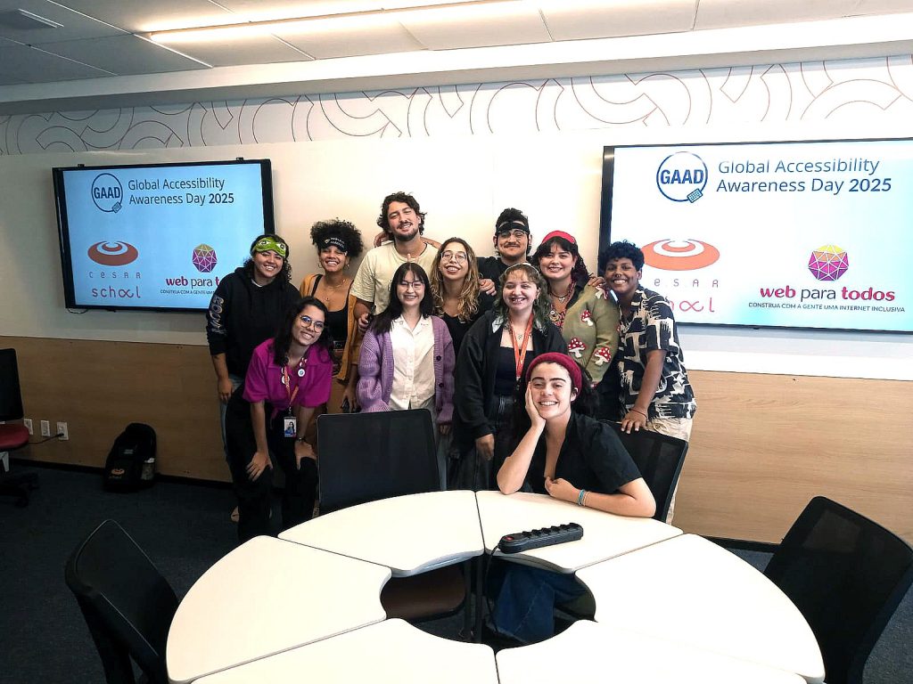 Foto de 11 estudantes universitários bem juntinhos e em frente a dois telões, onde está projetado nas telas os logotipos da CESAR School, do GAAD e do Web para Todos.