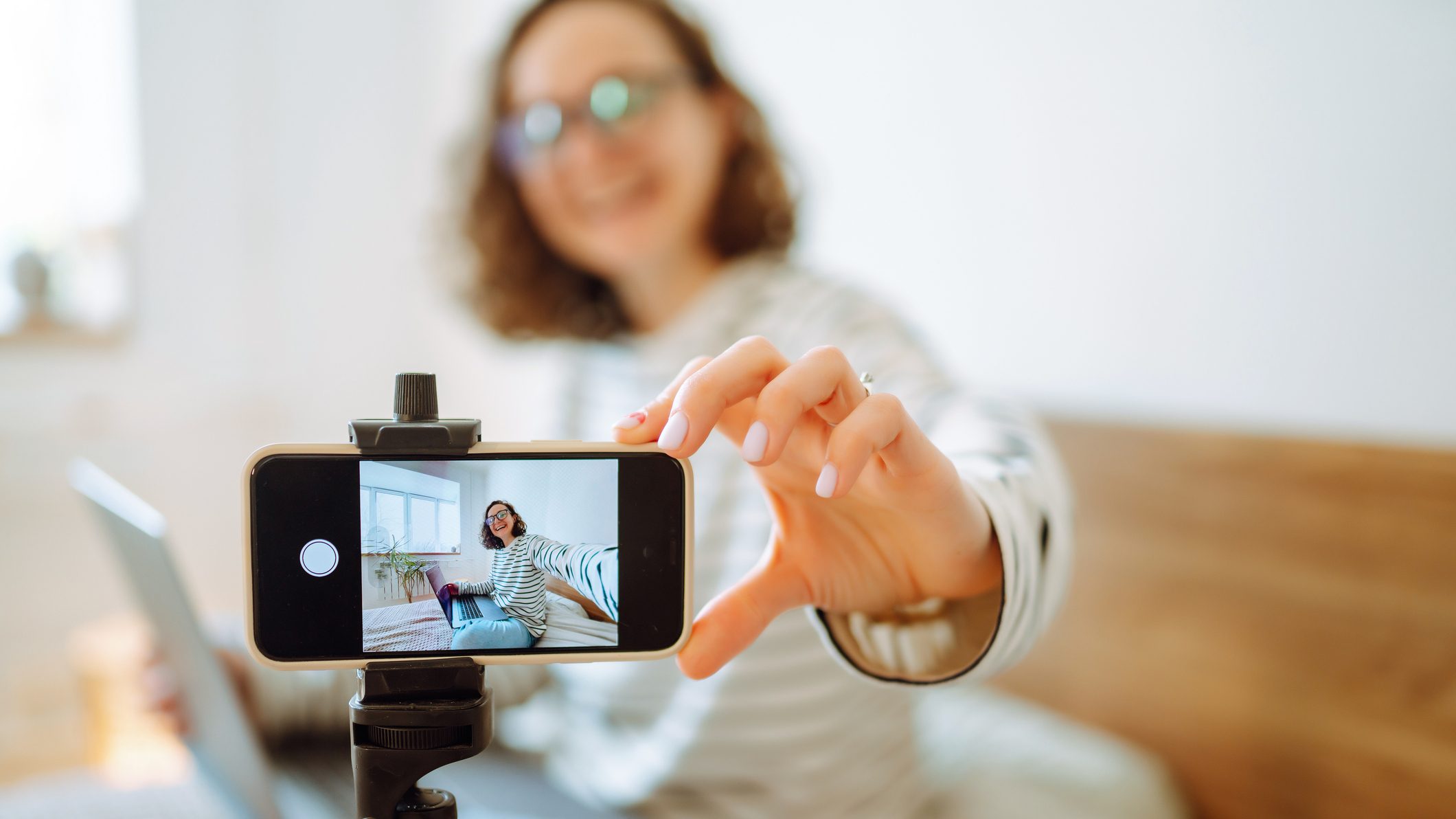 Foto de uma mulher segurando um smartphone montado em um tripé. Ela parece tirar uma selfie ou gravar um vídeo. Na tela do celular, vemos essa mesma pessoa sentada em uma cama, com um laptop sobre suas pernas. Ela sorri enquanto olha para a câmera.
