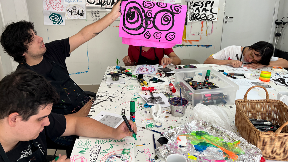 Foto de quatro pessoas da equipe do Estúdio QUE na sala de criação. Estão ao redor de uma mesa com tintas, canetinhas e papéis com muitas formas coloridas.