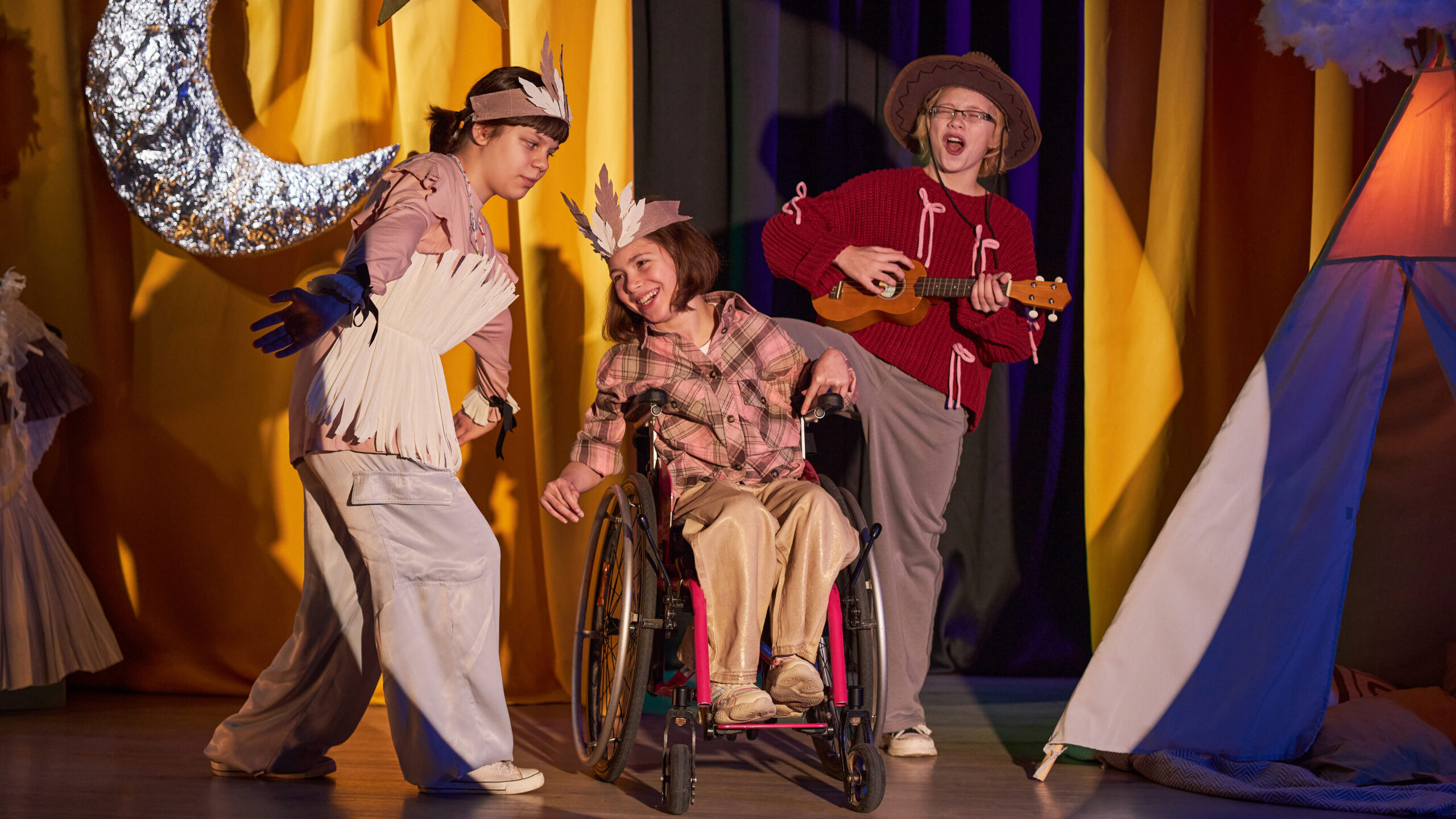 Foto de três crianças no palco durante apresentação de teatro. Elas estão fantasiadas e uma delas é cadeirante.