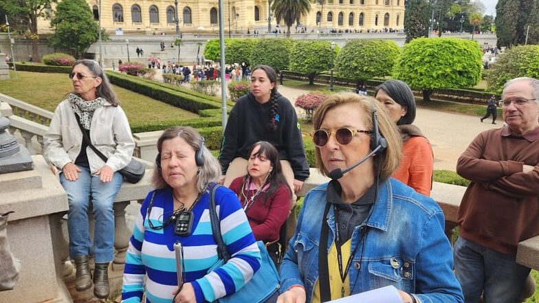 Foto de Lívia Motta e sua parceira de trabalho Cidinha Cardoso em área externa do Museu do Ipiranga, em São Paulo. Elas estão com fones de ouvido em meio a um grupo de pessoas.