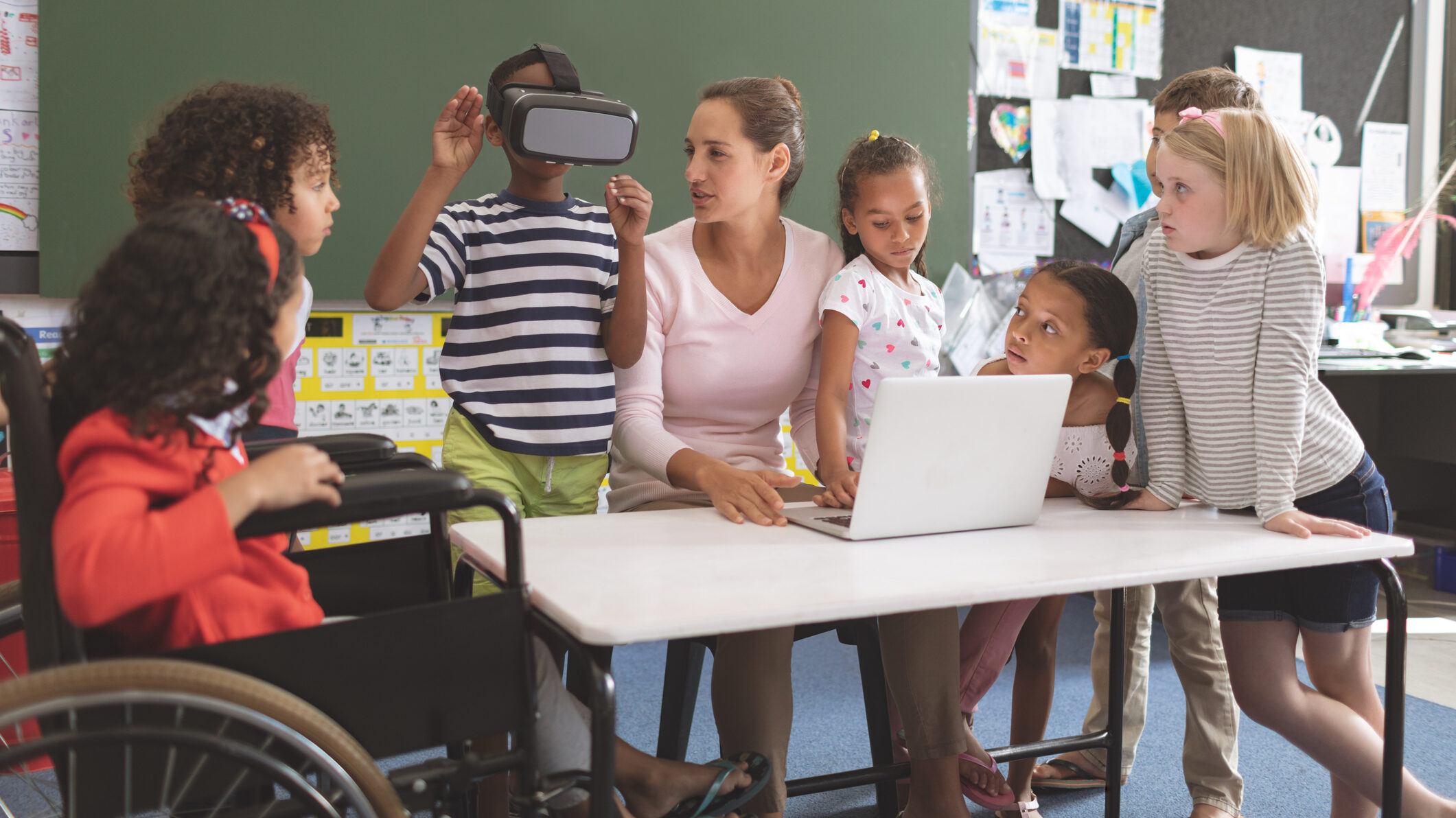 Foto de uma mulher rodeada por sete crianças, sendo uma cadeirante, em sala de aula. Uma delas usa óculos de realidade virtual enquanto as outras olham. Sobre a mesa há um notebook aberto.