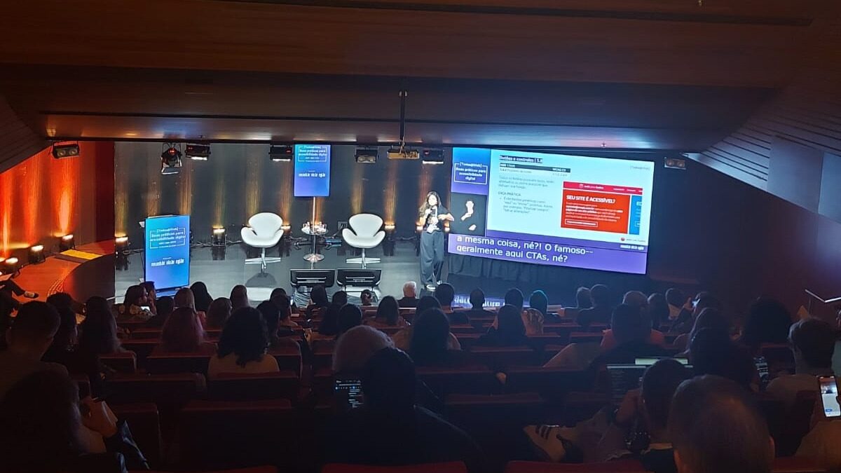 Foto do auditório onde aconteceu um dos eventos sobre a norma ABNT NBR 17225. Há muitas pessoas na plateia e a Simone Freire está no palco ao lado de um grande telão.