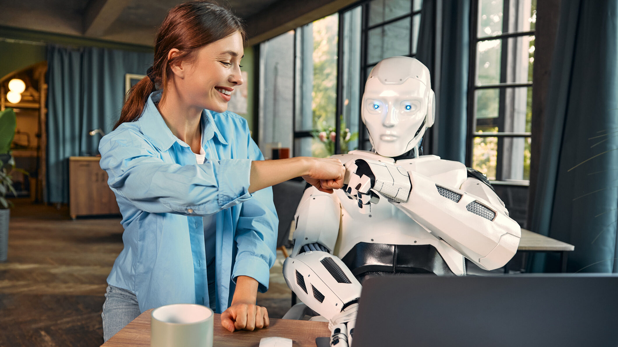 foto de um robô humanoide e uma jovem mulher sentados lado a lado em uma mesa, de frente a um notebook. Ambos estão se cumprimentando com punhos fechados (dando o que chamamos de "soquinho"), numa interação colaborativa. Estão em um ambiente interno com janelas grandes com cortinas azuis e plantas, compondo um cenário confortável e contemporâneo.