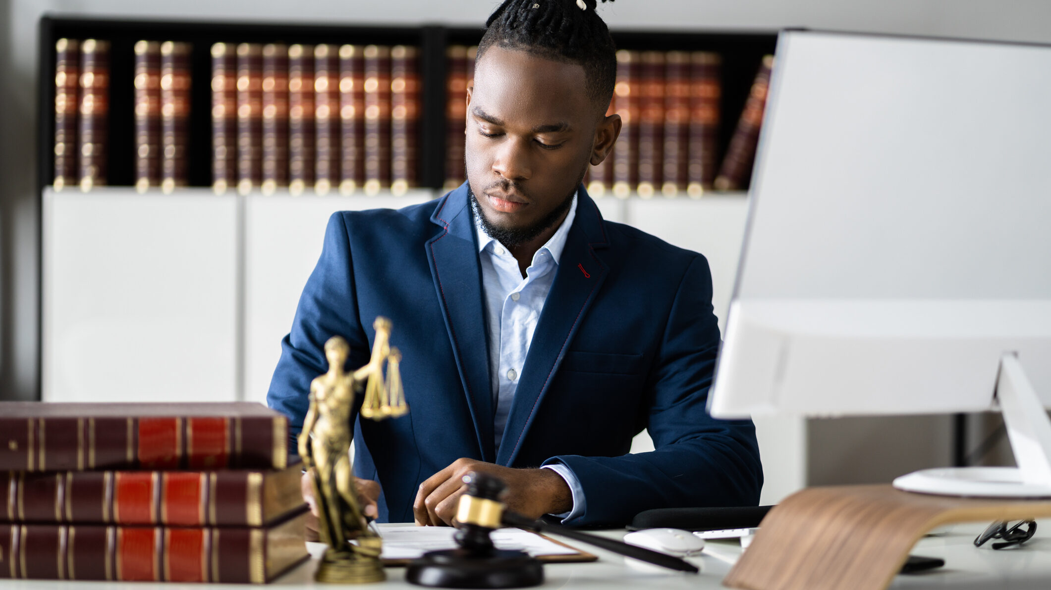 Foto de um rapaz negro com traje social sentado em uma mesa com computador, livros e objetos que remetem à Justiça, como o martelo utilizado em tribunais. Ele está sério e escreve em um caderno.