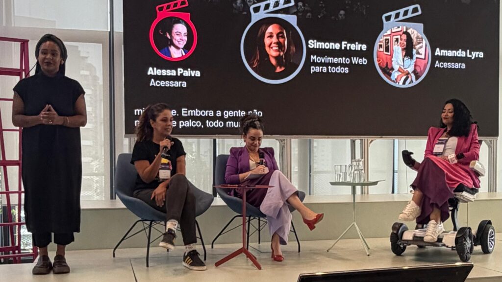 Foto do palco do evento de lançamento dos resultados da Pesquisa Nacional de Acessibilidade nos Cinemas. Sobre ele estão a intérprete de Libras, a mediadora Alessa Paiva, além das painelistas Simone Freire e Amanda Lyra. Atrás delas há um telão onde estão projetados o logotipo da pesquisa, fotos e nomes das participantes e a legenda em tempo real.