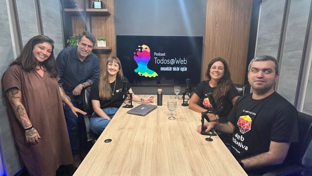 Foto de 5 pessoas ao redor de uma mesa retangular com microfones grandes, copos de água e notebook. Elas estão num ambiente fechado com paredes cinzas, móveis de madeira clara e, ao fundo, um monitor grande com o uma arte estilizada e o texto "Podcast Todos na Web. Ceweb.br, nic.br e cgi.br".
