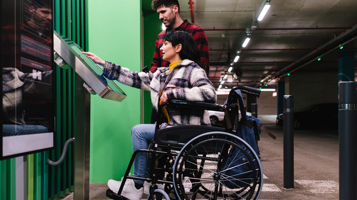 Foto de uma mulher em cadeira de rodas tocando o painel digital de um parquímetro acessível dentro de um estacionamento coberto. Ao lado dela há um homem em pé que observa a cena.