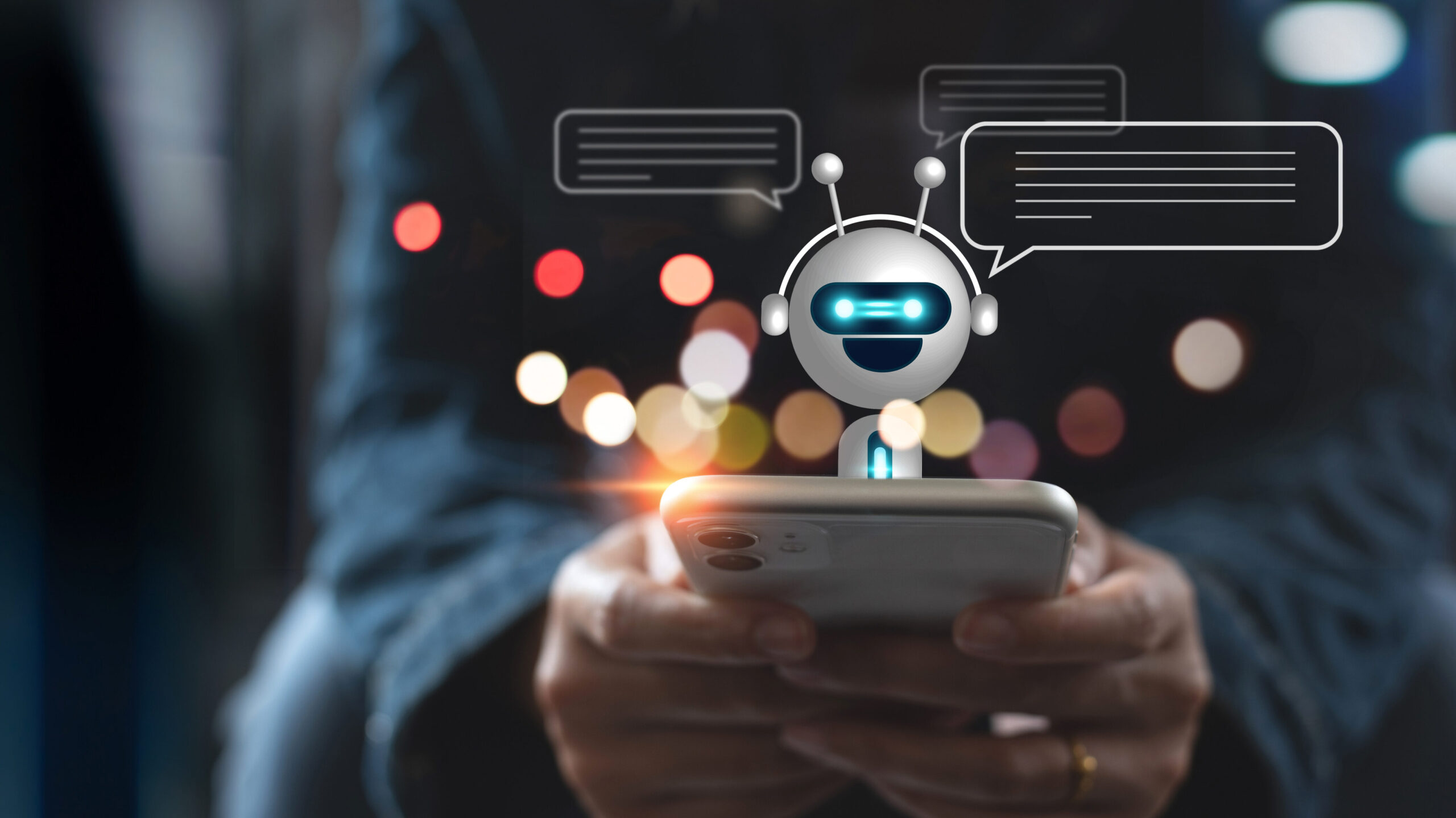 Foto artística de duas mãos humanas segurando um celular. Há um robô pequeno sorrindo sobre a tela com balões de diálogo próximos à sua cabeça. O fundo a foto está desfocado e há pontos de luzes coloridas. 