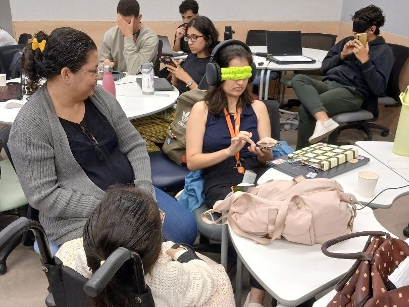 Foto de uma sala de aula com grandes mesas redondas. Ao redor delas, estudantes de Design usam dispositivos eletrônicos, fones de ouvido e vendas nos olhos para simular o uso de leitores de tela. Em primeiro plano, uma pessoa em cadeira de rodas acompanha a atividade.
