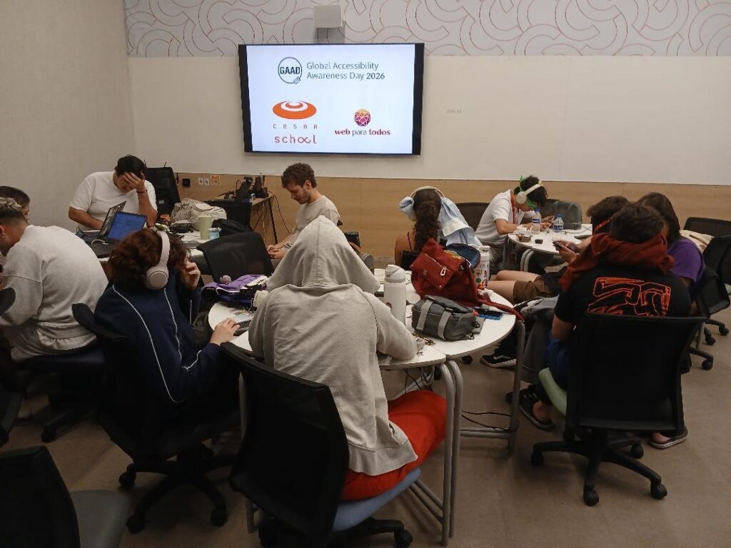 Foto de uma sala de aula com estudantes de Design reunidos em mesas redondas. De costas e de perfil, eles interagem com celulares sob simulação de acessibilidade, com uso de fones de ouvido, vendas e panos sobre os olhos. Ao fundo, uma tela de TV exibe os logos do "Global Accessibility Awareness Day 2026", "CESAR School" e "Movimento Web para Todos".