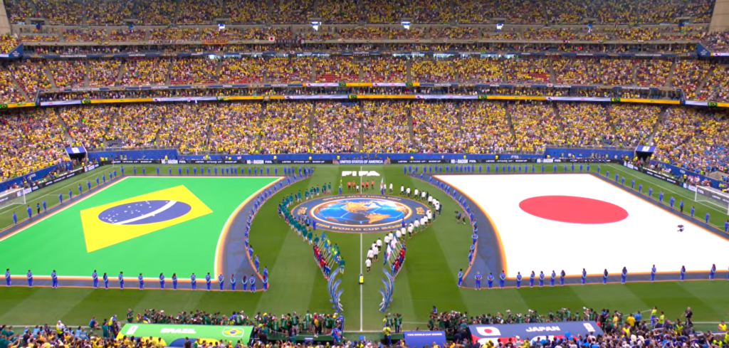 Foto aérea do gramado do NRG Stadium com as bandeiras gigantes do Brasil e do Japão estendidas antes do jogo. A arquibancada está lotada e os times estão em campo para a execução dos hinos nacionais.