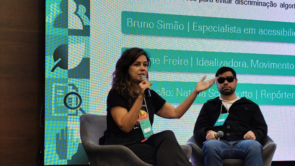 Foto de Simone Freire, sentada em uma poltrona no palco do evento, ao lado de outro palestrante do painel. Ela fala ao microfone, que segura com a mão direita, e ergue a mão esquerda para enfatizar a fala.