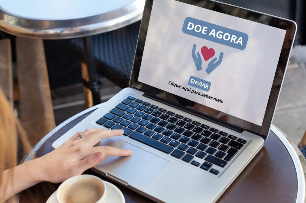 Foto de um laptop aberto sobre uma mesa redonda de café. A mão esquerda de uma pessoa está apoiada no touchpad do teclado. A tela exibe uma página de doação com o texto "DOE AGORA", um ícone de coração vermelho entre duas mãos azuis, o botão "ENVIAR" e a frase "Clique aqui para saber mais".