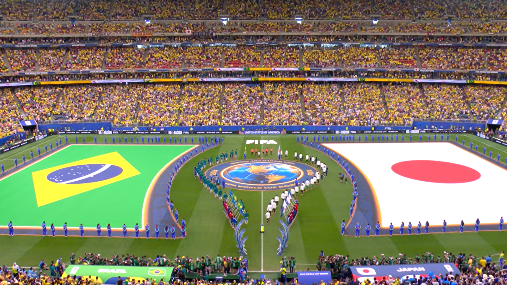 Foto aérea do gramado do NRG Stadium com as bandeiras gigantes do Brasil e do Japão estendidas antes do jogo. A arquibancada está lotada e os times estão em campo para a execução dos hinos nacionais.
