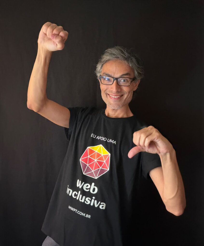 Alexandre Ohkawa sorri enquanto faz o sinal do número 9 em Libras com a mão esquerda e mantém a mão direita fechada em comemoração. Ele veste uma camiseta preta com a marca do Web para Todos e a frase 'Eu apoio uma web mais inclusiva'.