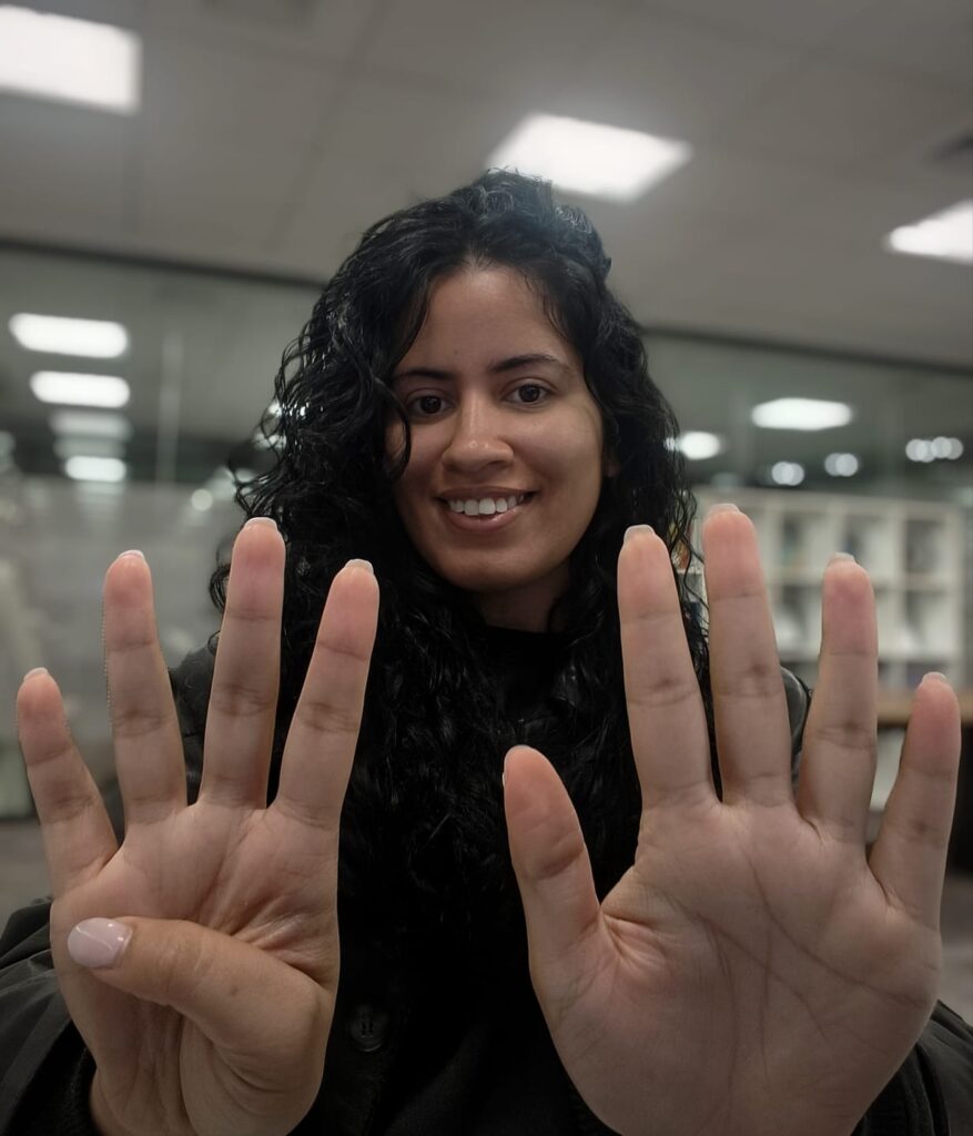 Amanda Marques sorri enquanto faz o número 4 com os dedos da mão direita e o número 5 com a mão esquerda, simbolizando o número 9.
