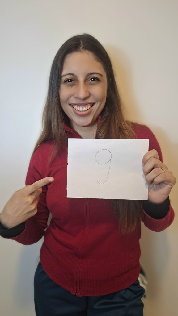 Ayna Rodrigues sorri enquanto segura com a mão esquerda uma folha de papel com o número 9 escrito, enquanto o indicador direito aponta para a folha.
